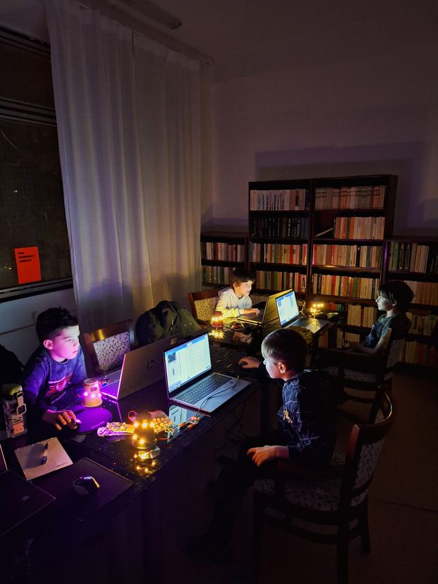 Deti na krúžku programovania sedia pri notebookoch v zatemnenej miestnosti, pred nimi svietia lampáše, ktoré samy postavili a naprogramovali na micro:bite