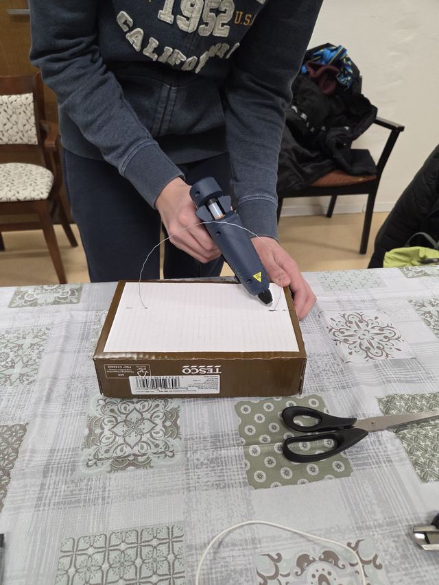 Dieťa lepí tavnou pištoľou drôt na kartónovú škatuľu, konštrukciu projektu si stavia samo